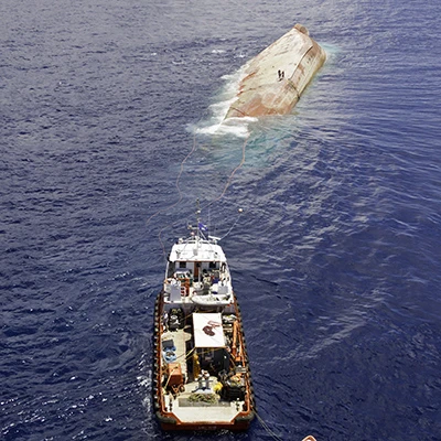Two people stand on the upturned hull of a sinking ship. Resolve Marine ships come to assist.